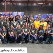 A group of people wearing matching shirts and some with gloves are posing inside a warehouse with stacked boxes. They appear to be participating in a volunteer event at the Los Angeles Regional Food Bank, dedicated to helping the community.