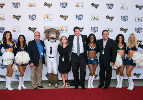 A group of people, including cheerleaders, a mascot, and men in suits, stand on a red carpet in front of a backdrop with logos for the Los Angeles Regional Food Bank and the Rams. Everyone is smiling and posing for the photo.