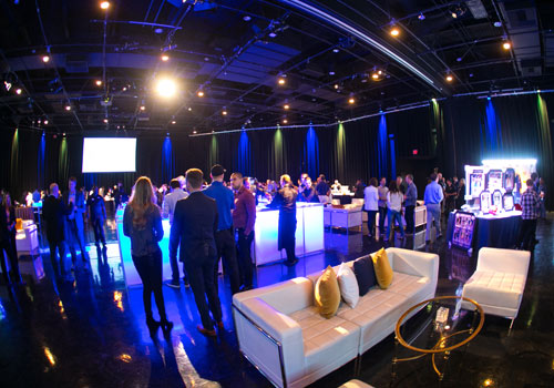 A modern event space buzzes with lively conversation as people gather around glowing tables, under soft overhead lighting. With white couches and yellow pillows, the room exudes warmth and vibrancy, echoing the community spirit of a Los Angeles Regional Food Bank event.