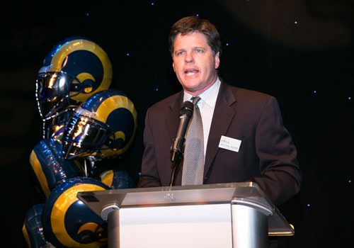A man in a suit speaks at a podium, representing the Los Angeles Regional Food Bank. Behind him, blue and yellow balloons shaped like helmets with a ram logo adorn the stage. The dark background is dotted with small lights, setting a hopeful tone for the event.