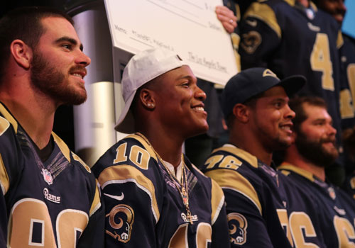 Four football players wearing navy and gold jerseys are seated in a row, smiling and looking to the right. One player sports a white cap. They appear to be at an event, possibly in support of the Los Angeles Regional Food Bank, with a blurred background setting the scene.