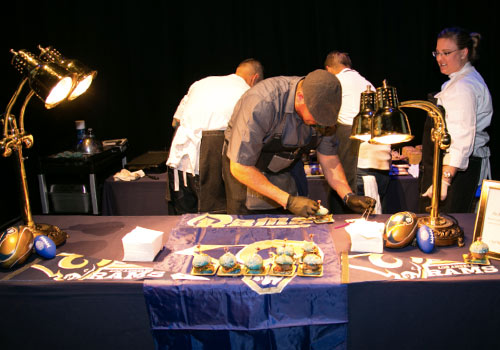 A chef prepares food on a table draped with a Rams-themed cloth, surrounded by others in white jackets at a charity event for the Los Angeles Regional Food Bank. Desserts and Rams-themed footballs are artfully displayed under warm lights, adding to the inviting atmosphere.