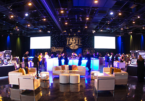 A dimly lit event hall with a modern setup featuring white couches and a central blue-lit bar buzzes with conversation. Guests mingle under the backdrop displaying "Taste of the NFL" amidst star-like lights, all while supporting the Los Angeles Regional Food Bank.