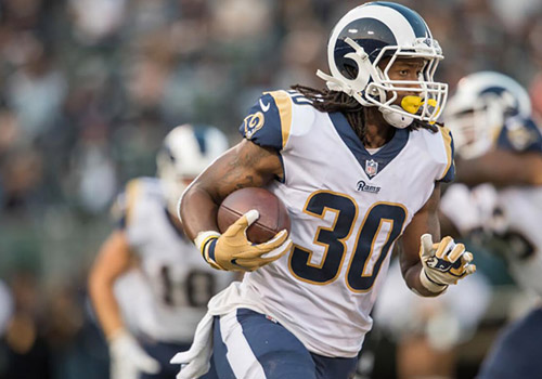 A football player wearing a white and blue uniform with number 30 runs with the ball, heading forward during a game. The logo on the helmet indicates he plays for the Rams. Teammates and an out-of-focus background suggest a stadium environment, possibly supporting the Los Angeles Regional Food Bank.