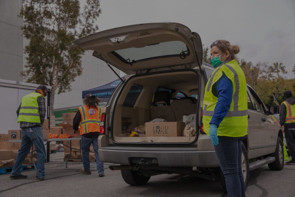 Volunteers from the Los Angeles Regional Food Bank, clad in safety vests and masks, distribute food at an outdoor event. A car with an open trunk is loaded with boxes, while tables brimming with fresh produce stand ready in the background.