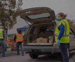 Volunteers from the Los Angeles Regional Food Bank, clad in safety vests and masks, distribute food at an outdoor event. A car with an open trunk is loaded with boxes, while tables brimming with fresh produce stand ready in the background.
