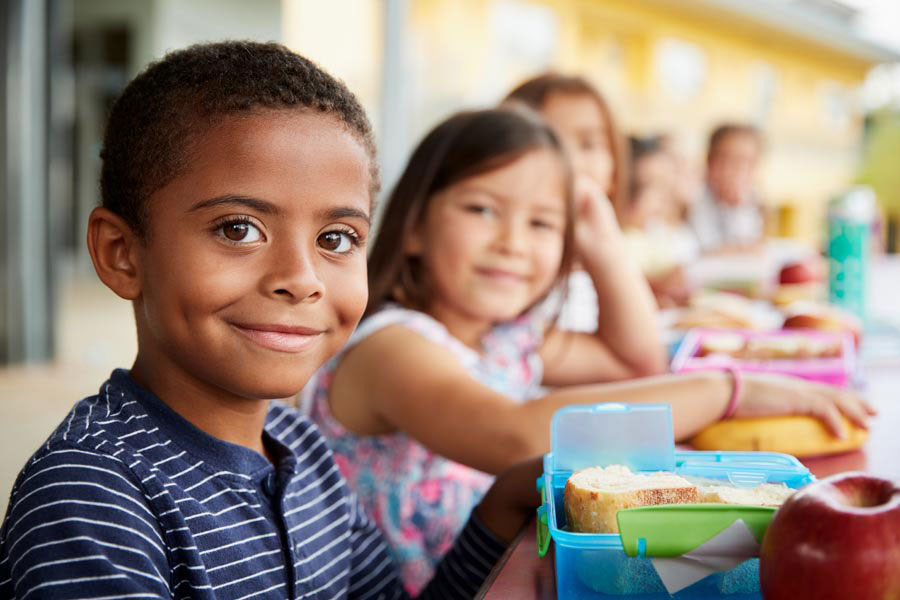 A group of children is sitting at a table, smiling while enjoying a meal from the Los Angeles Regional Food Bank. The focus is on a boy in the foreground with short hair and a striped shirt. Lunch boxes and food items are visible on the table, while the background remains slightly blurred.